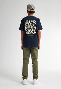 Navyblauwe katoenen t-shirt met een crèmekleurig graphic ontwerp op de achterkant, gecombineerd met groene broek en witte sneakers.