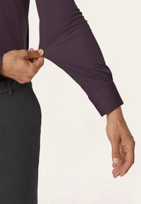 Langarmshirt in dunkellila mit glatter Textur und umschnittenem Ärmel. Eine Hand zieht den Ärmel leicht. 