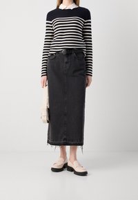 Kobieta w czarno-białym paskowanym swetrze, czarnej midi spódnicy z wysokim stanem i surowym wykończeniem, w beżowych mokasynach, trzymająca beżową torebkę.