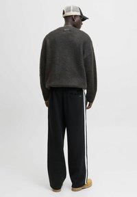 Mand iført sort sweater, sorte bukser med hvide striber, beige kasket og brune støvler, stående og vende væk mod en ensfarvet hvid baggrund.