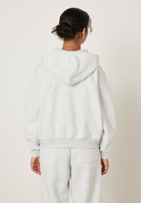 Šviesiai pilkas gobtuvu megztinis laisvo kirpimo, su ribuotais rankogaliais ir apačia, pagamintas iš minkštos tekstūruotos medžiagos.