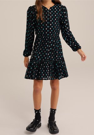 Dziewczyna ubrana w czarną sukienkę z długim rękawem w niebiesko-biały kwiatowy wzór, do tego czarne skarpetki ozdobione perłami oraz czarne buty na pasku typu T.