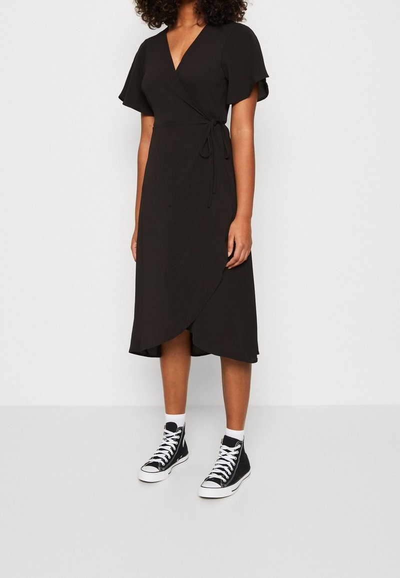 Žena v černých zavinovacích šatech s volnými rukávy v kombinaci s bílými ponožkami a černými vysokými teniskami stojí před jednobarevným pozadím.