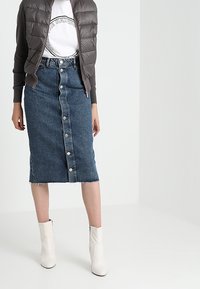 Falda de denim con cierre delantero de botones, dobladillo deshilachado y forma de lápiz, combinada con una camisa blanca y un chaleco acolchado gris; modelo con botas blancas hasta el tobillo.