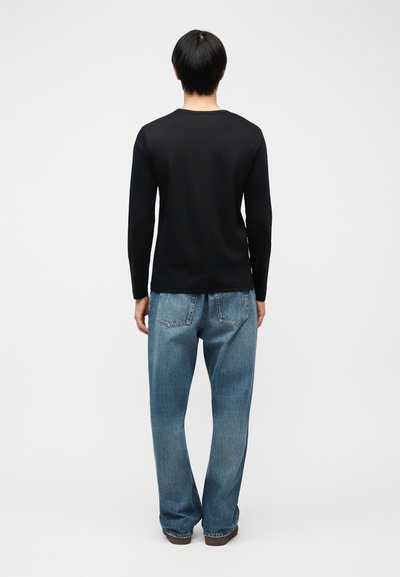 Άτομο στέκεται έχοντας την πλάτη του προς τα εμπρός, φορώντας μαύρο μακρυμάνικο πουκάμισο, άνετο μπλε τζιν και καφέ παπούτσια, μπροστά σε ένα απλό ανοιχτόχρωμο φόντο.