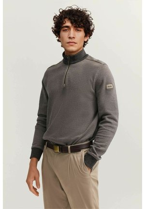 Junger Mann mit lockigem Haar, trägt einen grauen Viertel-Reißverschluss-Pullover und beige Hose mit einem dunkelbraunen Gürtel, steht mit einer Hand in der Tasche.