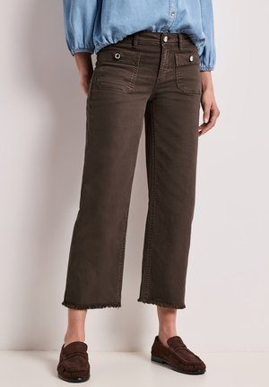 Person iført cropped mørkebrune bukser med forlommer og frynser i kanterne, kombineret med mørkebrune loafers og en lyseblå skjorte.