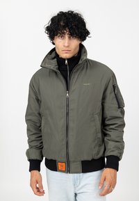 Veste bomber vert olive avec col haut, fermeture éclair à l'avant, poche latérale et étiquette orange sur l'ourlet. Poignets et taille côtelés.