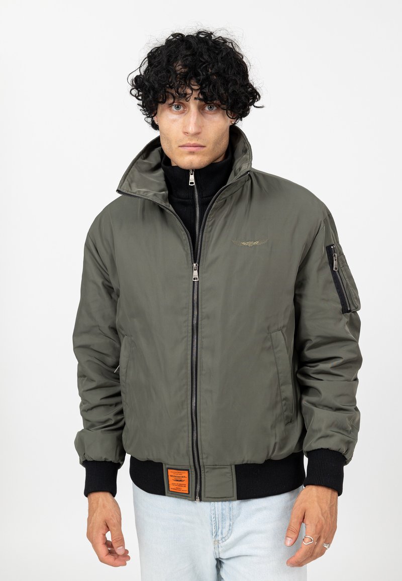 Veste bomber vert olive avec col haut, fermeture éclair à l'avant, poche latérale et étiquette orange sur l'ourlet. Poignets et taille côtelés.