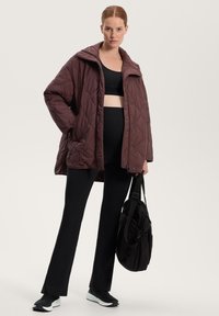 Gequilted, bordeaux jacket met een hoge kraag, gecombineerd met een zwarte crop top en wijde broek. Een zwarte tas wordt in één hand vastgehouden.