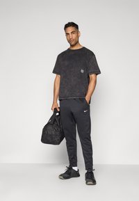 T-shirt en coton noir usé avec un petit logo sur la poitrine, associé à un pantalon de survêtement Nike gris foncé et des chaussures de sport noires. Tient un sac de sport noir.