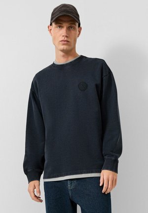 Junger Mann trägt ein dunkelblaues Sweatshirt, schwarze Kappe und Jeans und steht vor einem schlichten hellgrauen Hintergrund.