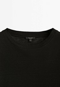 T-shirt noir à col rond et manches courtes avec une étiquette de taille visible indiquant une taille moyenne pour l’UE, les États-Unis et le Mexique.