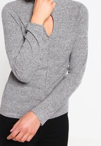 Grå stickad tröja med långa ärmar och V-ringning. Har ett strukturerat tyg med en figurnära design. Svarta byxor syns nedanför.