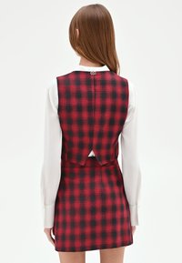 Vestito a quadri rosso e nero con corpetto aderente e gonna svasata. Presenta un'apertura posteriore con bottone ed è abbinato a una camicia bianca a maniche lunghe.