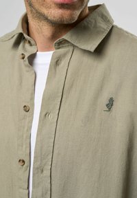 Camisa de lino verde olivo con botones en la parte delantera, botones marrones y un pequeño logotipo bordado en el lado izquierdo del pecho. Camiseta blanca debajo.