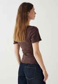 Hnedý rebrovaný top s krátkymi rukávmi a vlnitým lemom, skombinovaný s tmavými džínsami, z bočného pohľadu. Hladká textúra, priliehavý strih.