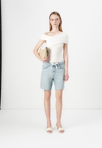 Bijela off-shoulder majica, svijetloplave traper šortse, bež natikače, drži torbicu od teksturirane krem boje. Jednostavne linije i opušteni silueta.