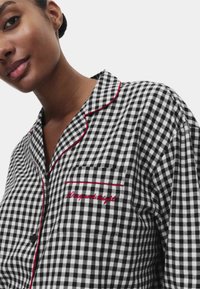 Sort-hvidt gingham-mønsteret pyjamasoverdel med røde piping og broderet tekst "Daisy and Bright." Har en brystlomme og knaplukning.