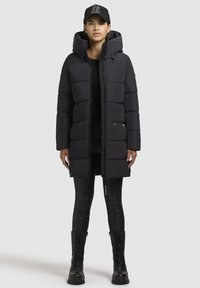 Zwarte pufferjas met een capuchon, voorzien van gequiltte segmenten, zijzakken en een volledige rits. Gepaard met zwarte leggings en veterschoenen.