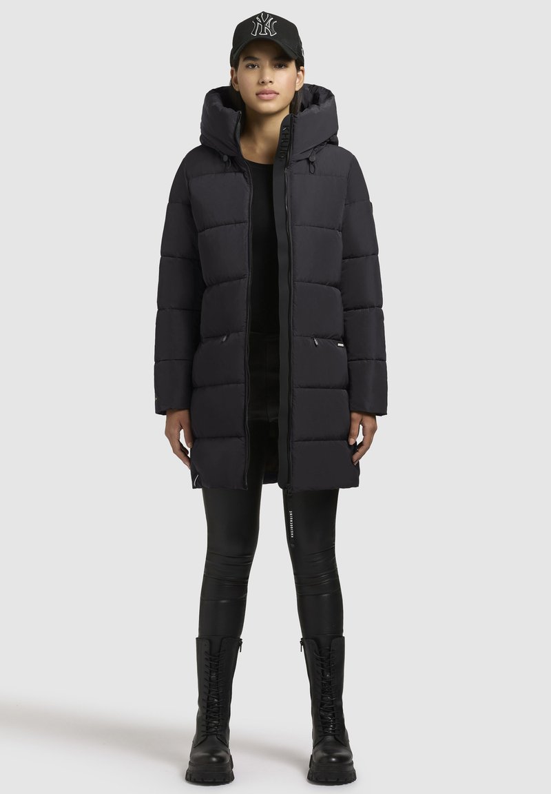 Zwarte pufferjas met een capuchon, voorzien van gequiltte segmenten, zijzakken en een volledige rits. Gepaard met zwarte leggings en veterschoenen.