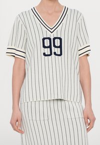 Bílé tričko ve stylu baseballu s pruhy, výstřihem do V a krátkými rukávy. Má tmavě modré pruhy a číslo "99" na přední straně. Měkký, texturovaný pletený materiál.