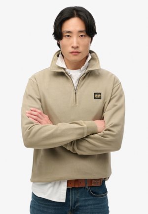 Sudadera beige de medio cierre fabricada en suave algodón, con puños acanalados y un parche de logo en el pecho. Lleva puesta sobre una camisa blanca con jeans azules.