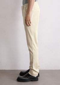 Persoon in beige slim-fit broek en zwarte leren instappers, zijwaarts staand tegen een licht gestructureerde muur.