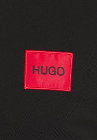 Patch rectangulaire rouge en tissu avec le texte noir "HUGO" cousu sur un fond en tissu noir texturé.