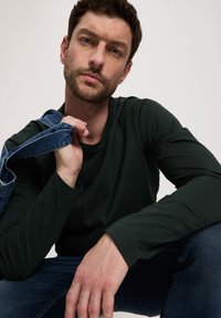 Ciemnozielona koszula z długim rękawem i okrągłym dekoltem, w połączeniu z niebieskimi dżinsami. Osoba trzyma dżinsowe ogrodniczki na ramieniu.