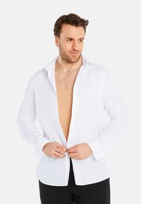 White button-up shirt with long sleeves, a spread collar, and a smooth texture. Worn open over a tan undershirt. Black trousers visible.
