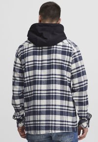 Camicia a quadri in blu navy e bianco con cappuccio blu navy. Presenta maniche lunghe e polsini con bottone. Il materiale sembra morbido con una finitura testurizzata.