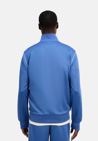 Blaue Trainingsjacke aus glattem Stoff mit hohem Kragen, gerippten Bündchen und weißen Paspelierungen an den Schultern.