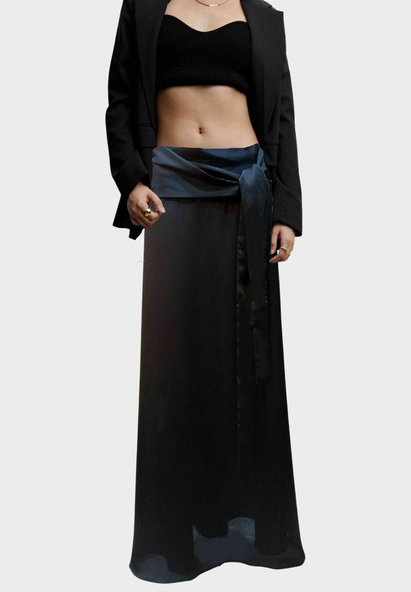Falda maxi negra con una cinturilla de satén, que presenta un detalle atado. Combinada con una blusa negra ajustada y una chaqueta entallada. Textura suave.