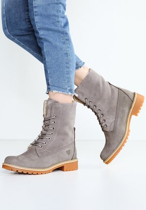 Bottes en daim gris à lacets avec semelles en caoutchouc marron portées avec un jean bleu coupé sur fond blanc.