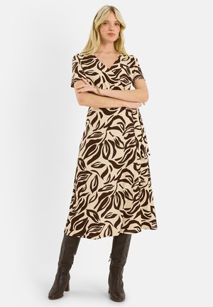 Mujer rubia con los brazos cruzados, vestida con un vestido cruzado beige con un estampado de hojas marrones y botas marrón oscuro hasta la rodilla, sobre un fondo blanco.