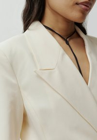 Beige maßgeschneiderter Blazer mit Reverskragen, der über einen tiefen V-Ausschnitt und einen schwarzen Kordelzugakzent um den Hals verfügt. Glatte Textur.