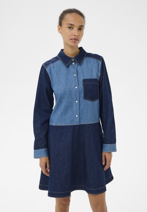 Kvinde iført en langærmet, knap-op denimkjole med lyse og mørkeblå farveblokke og en mørkeblå lomme på brystet.