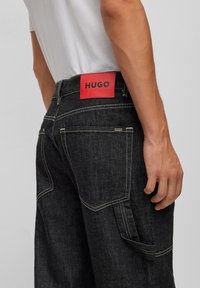 Černé džíny mají velkou červenou nášivku s nápisem "HUGO" na pásku, kontrastní švy a boční kapsu.