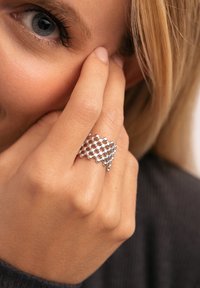 Anillo de plata con un diseño texturizado y trenzado que presenta pequeños acentos esféricos. La mano de la modelo toca suavemente su rostro, resaltando el anillo.