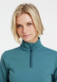 Pullover teal con colletto alto, zip quarti, texture liscia, con sottile branding sul petto e cuciture a contrasto.