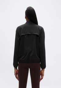 Άτομο που φαίνεται από πίσω φορώντας μια  εφαρμοστή μαύρη ζακέτα με λεπτομέρεια αερισμού στο πίσω μέρος και σκούρα καφέ κολάν σε απλό φόντο.