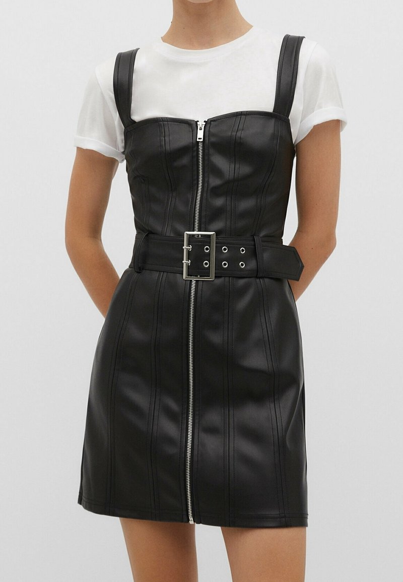 Femme portant une mini-robe noire en simili cuir avec une fermeture éclair à l'avant et une large ceinture sur un t-shirt blanc à manches courtes, debout devant un fond uni.