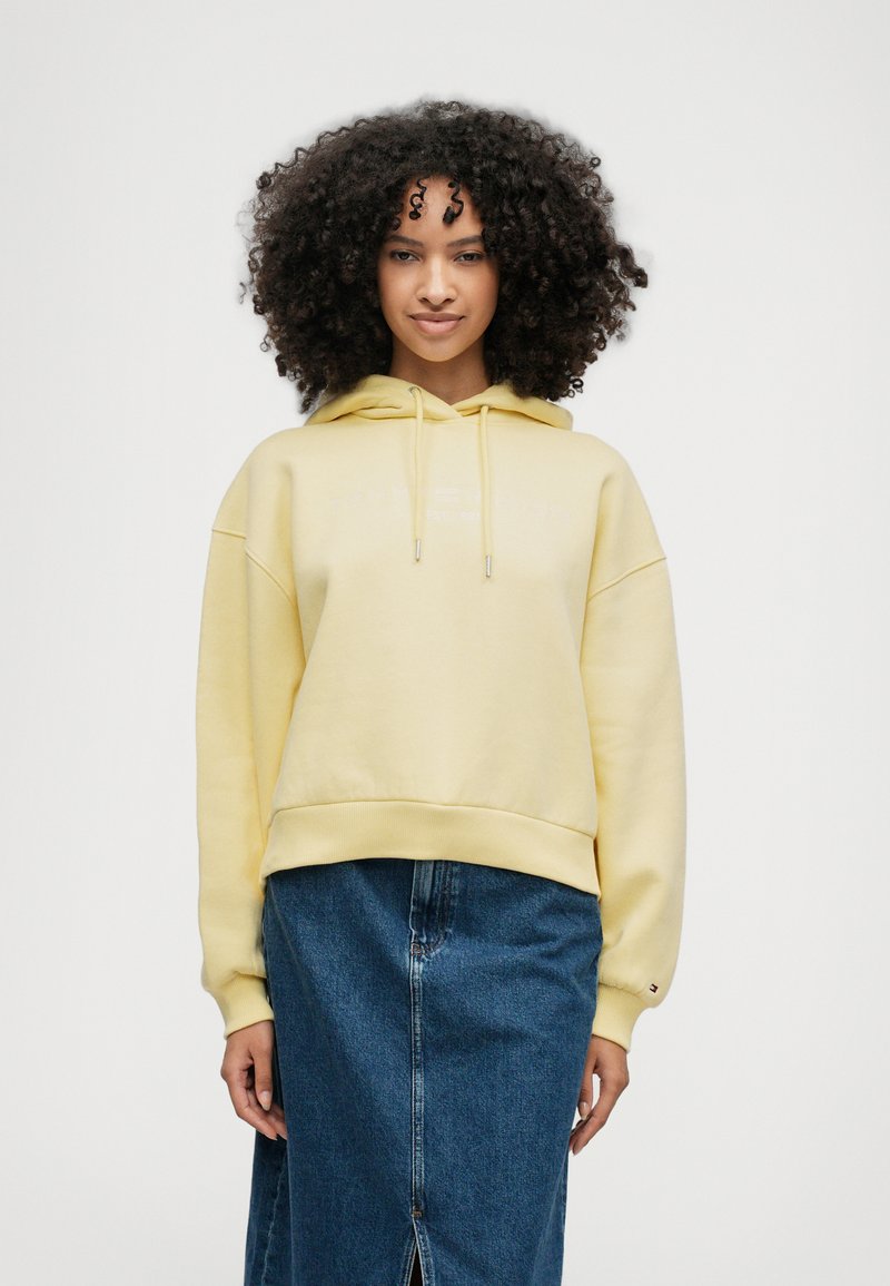 Κίτρινη βαμβακερή φούτερ με κουκούλα, σε χάλαρη εφαρμογή, με κορδόνι και ριμπ μανσέτες, συνδυασμένη με τζιν φούστα με μπροστινή σχισμή.