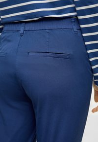 Blaue Hosen aus glattem Stoff, mit einem elastischen Bund, einer Gesäßtasche und subtilen Glanz, kombiniert mit einem gestreiften blauen Hemd.