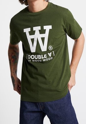 Mężczyzna ubrany w oliwkową koszulkę z dużym białym logo "WW" i tekstem "Double A by Wood Wood", w parze z ciemnoniebieskimi dżinsami, tatuaże widoczne na lewej ręce.