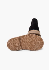 Chaussure slip-on en suede noir avec une semelle en caoutchouc brun texturé. La chaussure présente un design sans couture et un bout arrondi pour un look minimaliste.
