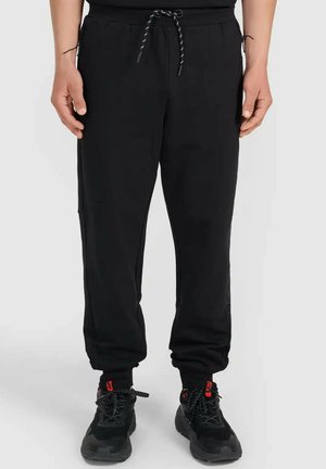 Mężczyzna w czarnych spodniach dresowych z sznurkiem w talii i czarnych butach sportowych stoi na białej podłodze.