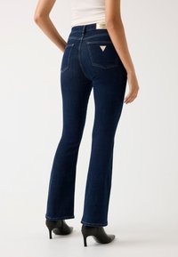 Jean flare bleu marine en denim, avec deux poches arrière affichant un logo en triangle. L'ourlet est légèrement effiloché. Porté avec des bottes à talons noires.