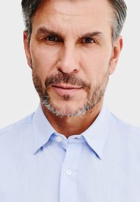 Jasnoniebieska koszula zapinana na guziki, wykonana z lekkiego materiału, z klasycznym kołnierzykiem i starannie wyprasowana, noszona przez mężczyznę z brodą.